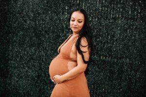 Pregnant woman in a rust orange dress posing outdoors showing what to wear to a maternity photo shoot.