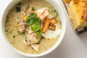 Bowl of creamy slow cooker white chicken chili topped with jalapeños and tortilla strips beside cornbread.